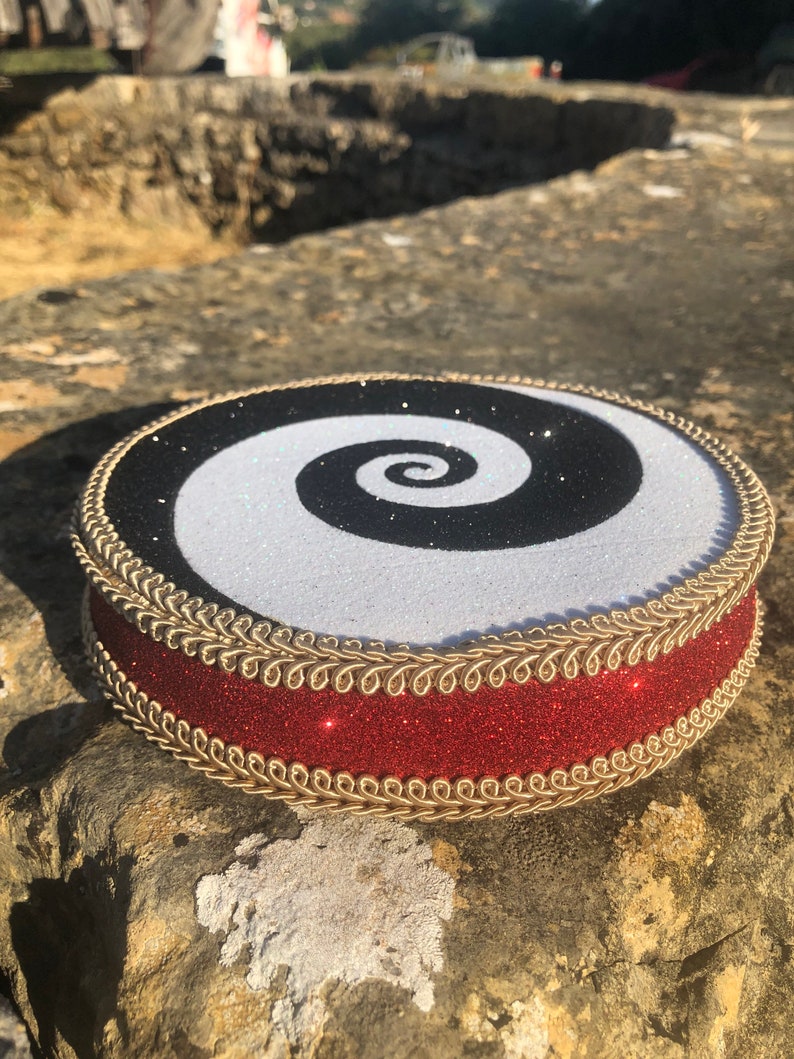 May include: A round cake topper with a black and white spiral design. The topper is decorated with gold trim and a red glitter ribbon.