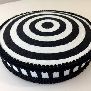 Puede incluir: Un soporte para tartas redondo, blanco y negro con un dise&ntilde;o de c&iacute;rculos conc&eacute;ntricos. El soporte est&aacute; decorado con una cinta negra y una cinta a rayas blancas y negras.