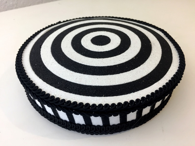 Puede incluir: Un soporte para tartas redondo, blanco y negro con un dise&ntilde;o de c&iacute;rculos conc&eacute;ntricos. El soporte est&aacute; decorado con una cinta negra y una cinta a rayas blancas y negras.