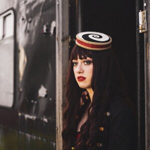 May include: A woman with long dark hair wears a black and white spiral patterned hat with a red band. She is wearing a black jacket with gold trim and a red dress.