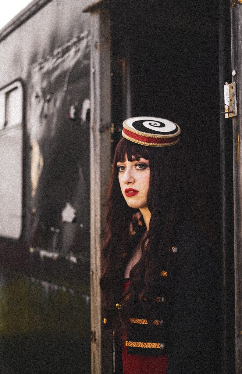 May include: A woman with long dark hair wears a black and white spiral patterned hat with a red band. She is wearing a black jacket with gold trim and a red dress.