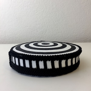 Puede incluir: Un coj&iacute;n circular blanco y negro con un patr&oacute;n de c&iacute;rculos conc&eacute;ntricos. El coj&iacute;n tiene un ribete a rayas blanco y negro.
