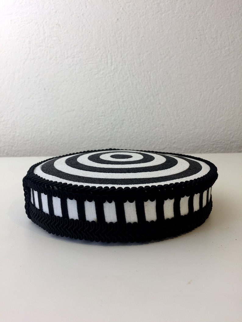 Puede incluir: Un coj&iacute;n circular blanco y negro con un patr&oacute;n de c&iacute;rculos conc&eacute;ntricos. El coj&iacute;n tiene un ribete a rayas blanco y negro.