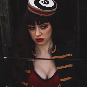May include: A woman wearing a black and white spiral patterned hat with a red band, a black jacket with gold trim, and a red top with gold rope accents.