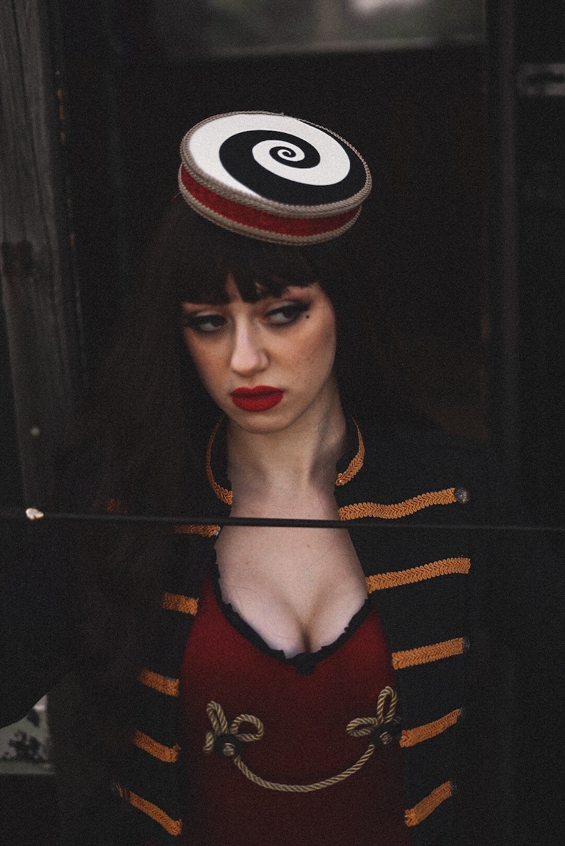 May include: A woman wearing a black and white spiral patterned hat with a red band, a black jacket with gold trim, and a red top with gold rope accents.