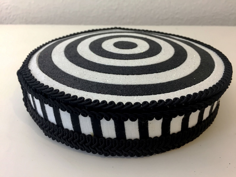 Puede incluir: Soporte para pastel redondo en blanco y negro con una cinta rayada en blanco y negro y un ribete trenzado negro. La parte superior del soporte tiene un dise&ntilde;o de c&iacute;rculos conc&eacute;ntricos en blanco y negro.
