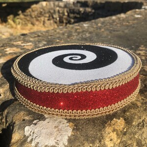 May include: A round, black and white spiral design cake topper with a red glitter ribbon and gold trim.