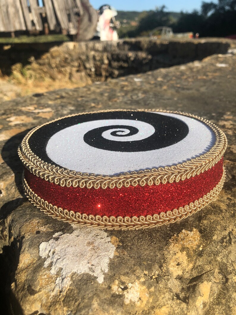 May include: A round, black and white spiral design cake topper with a red glitter ribbon and gold trim.
