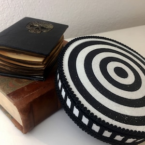 Puede incluir: Un coj&iacute;n redondo de color blanco y negro con un patr&oacute;n de espiral y un borde a rayas blanco y negro. El coj&iacute;n est&aacute; encima de una pila de dos libros. El libro de arriba es negro con un dise&ntilde;o de calavera dorada.