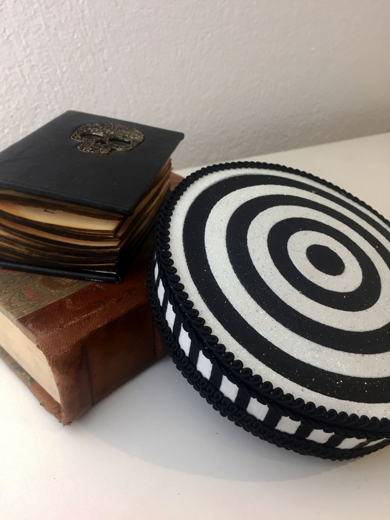 Puede incluir: Un coj&iacute;n redondo de color blanco y negro con un patr&oacute;n de espiral y un borde a rayas blanco y negro. El coj&iacute;n est&aacute; encima de una pila de dos libros. El libro de arriba es negro con un dise&ntilde;o de calavera dorada.