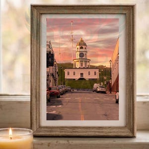 Puede incluir: Una fotografía enmarcada del reloj de Halifax al atardecer, que muestra una estructura blanca con una torre de reloj al final de una calle con coches aparcados, bajo un cielo rosa y naranja vibrante, presentada en un marco de madera clara.