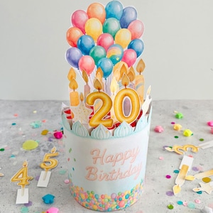 Könnte beinhalten: Papier-Geburtstagskuchen-Dekoration mit hellblauem Sockel, verziert mit bunten Konfetti und den Worten "Happy Birthday". Es zeigt eine "20" Kerze und eine Ansammlung von Ballons in verschiedenen Farben. Zusätzliche Zahlenkerzen und sternförmige Konfetti sind verstreut.