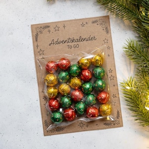 Puede incluir: Calendario de Adviento con bombones envueltos en papel dorado, rojo y verde. Los bombones están en una bolsa transparente, adheridos a una tarjeta marrón con el texto "Adventskalender TO GO" y adornos.