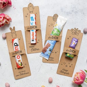 Puede incluir: Una variedad de golosinas de Pascua, incluyendo barras de chocolate y caramelos, presentadas en tarjetas decorativas de papel kraft. Las tarjetas presentan ilustraciones de un pollito, un conejito y una mariposa, con las palabras "für DICH" y "Frohe Ostern".