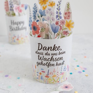 Op de afbeelding: Een witte papieren beker met een bloemenontwerp en de tekst "Danke, dass du uns beim Wachsen geholfen hast." Kleurrijke papieren bloemen zijn in de beker gerangschikt. Een tweede beker met "Happy Birthday" staat op de achtergrond.