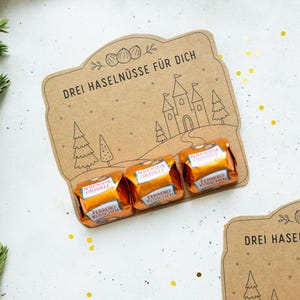 Könnte beinhalten: Eine braune Pappkarte mit dem Text DREI HASELNÜSSE FÜR DICH, einer Strichzeichnung eines Schlosses und Bäumen sowie drei Ferrero Küsschen in orangefarbenen Folienverpackungen, die am unteren Rand der Karte befestigt sind.