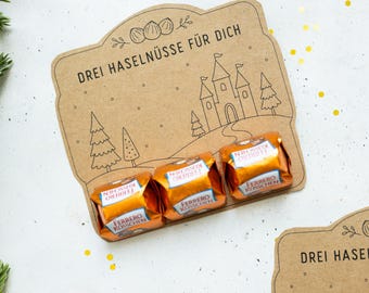 以下が含まれることがあります： DREI HASELNÜSSE FÜR DICHという文字、城と木の線画が描かれた茶色の厚紙カード。カードの下端には、オレンジ色のホイルで包まれた3つのフェレロ・キュッヒェンチョコレートが取り付けられています。