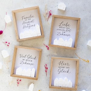 May include: Four square wooden shadow boxes containing white house-shaped figurines and printed text. The text reads: Familie ist die Heimat des Herzens, Liebe macht aus einem Ort ein Zuhause, Viel Glück im neuen Zuhause, and Hier wohnt das Glück.