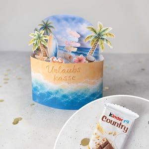 Peut inclure: Un récipient cylindrique avec un habillage sur le thème de la plage, illustré de palmiers, d'une planche de surf et d'un panneau, portant l'inscription Urlaubs kasse. Une barre chocolatée Kinder Country est placée sur une base circulaire blanche.