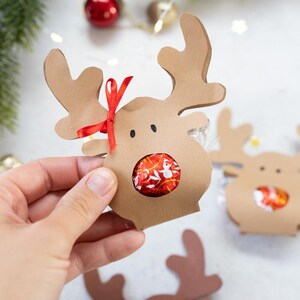 Puede incluir: Cajas de regalo con forma de reno marr&oacute;n con cinta roja y un caramelo envuelto en rojo y blanco en el centro. Las cajas tienen cuernos y ojos recortados. Embalaje de regalo navide&ntilde;o.