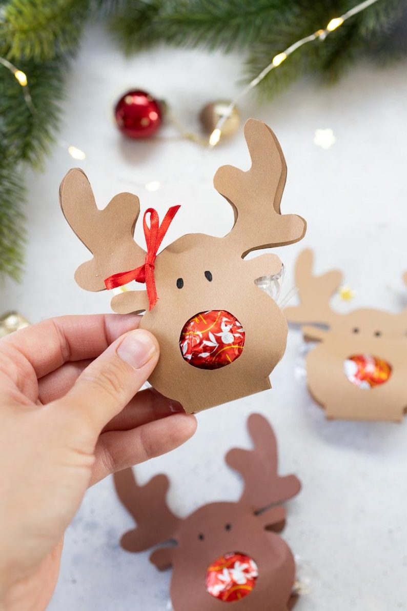 Puede incluir: Cajas de regalo con forma de reno marr&oacute;n con cinta roja y un caramelo envuelto en rojo y blanco en el centro. Las cajas tienen cuernos y ojos recortados. Embalaje de regalo navide&ntilde;o.