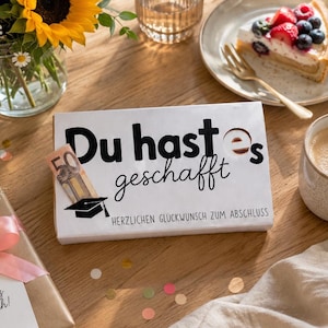 Peut inclure: Une boîte cadeau rectangulaire blanche avec le texte noir 'Du hast es geschafft' et 'HERZLICHEN GLÜCKWUNSCH ZUM ABSCHLUSS', un graphique de toque de diplômé et un billet de cinquante euros inséré sur le côté du couvercle.