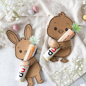 Op de afbeelding: Paasdecoraties met een konijn en een kuiken van bruin karton, elk met een chocoladereep in de vorm van een wortel. Op de chocoladerepen staat het woord "du". De achtergrond bevat paaseieren.