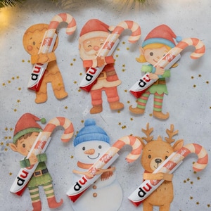 Peut inclure: Diverses découpes en papier sur le thème de Noël, dont un bonhomme en pain d'épices, le Père Noël, un elfe, un bonhomme de neige et un renne. Chaque personnage tient un sucre d'orge avec du texte. Les cannes sont rayées rouge et blanc.