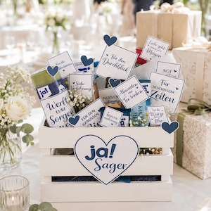 Puede incluir: Una caja de madera blanca llena de varios artículos pequeños y tarjetas con texto en alemán, incluyendo frases como "ja! Sager," "Werft Eure Sorgen weg," y "Für den Durchblick." La caja está decorada con recortes en forma de corazón azul oscuro.