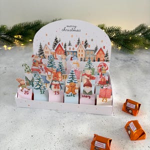 Könnte beinhalten: Ein weihnachtlicher Adventskalender aus Pappe mit einer festlichen Dorfillustration, nummerierten Türchen mit Weihnachtsmotiven und dem Text merry christmas. Drei orangefarbene Ferrero Küsschen liegen vor dem Kalender.