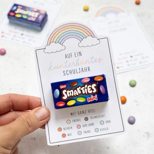Könnte beinhalten: Eine weiße Karte mit einer bunten Regenbogenillustration oben und dem Text AUF EIN kunterbuntes SCHULJAHR. Eine kleine blaue Schachtel Smarties mini ist in der Mitte befestigt, darunter eine Liste deutscher Wörter wie ENERGIE, FREUNDSCHAFT, NEUGIER, GUTE LAUNE, SPAB, MUT, FREUDE und GEDULD.