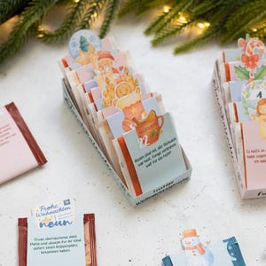 Könnte beinhalten: Eine Sammlung kleiner rechteckiger Tütchen in einer Schachtel, die jeweils mit bunten Weihnachtsmotiven und deutschem Text bedruckt sind. Die Tütchen zeigen festliche Grafiken wie einen Schneemann, Lebkuchen und Zuckerstangen sowie aufgedruckte Zahlen und Sprüche.