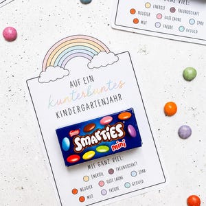 Könnte beinhalten: Eine weiße Karte mit einem Regenbogen und Wolken oben, darauf der Text AUF EIN kunterbuntes KINDERGARTENJAHR und MIT GANZ VIEL sowie eine Liste von Begriffen mit farbigen Kreisen. Eine blaue Schachtel Smarties mini ist auf der Karte befestigt.