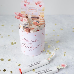 Könnte beinhalten: Eine weiße Pop-up-Hochzeitskarte mit einem Auto, Kuchen und Herzdekorationen. Auf der Karte steht "Alles Liebe zur Hochzeit" in roter Schrift. Im Vordergrund liegen zwei weiße Papierröhren mit Text und Illustrationen.