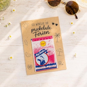 Op de afbeelding: Een rechthoekige kaart van kraftpapier met de tekst 'ICH WÜNSCHE DIR prickelnde Ferien' en kleine zwarte lijntekeningen van zomerse iconen. Een zakje Ahoj-Brause framboos bruispoeder is in het midden van de kaart bevestigd.