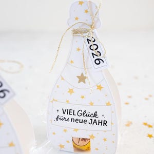 Puede incluir: Una caja de regalo blanca con forma de botella de champ&aacute;n, adornada con estrellas doradas y el texto "2026" en una etiqueta. La caja tiene una cuerda dorada y el texto alem&aacute;n "VIEL Gl&uuml;ck f&uuml;rs neue JAHR".