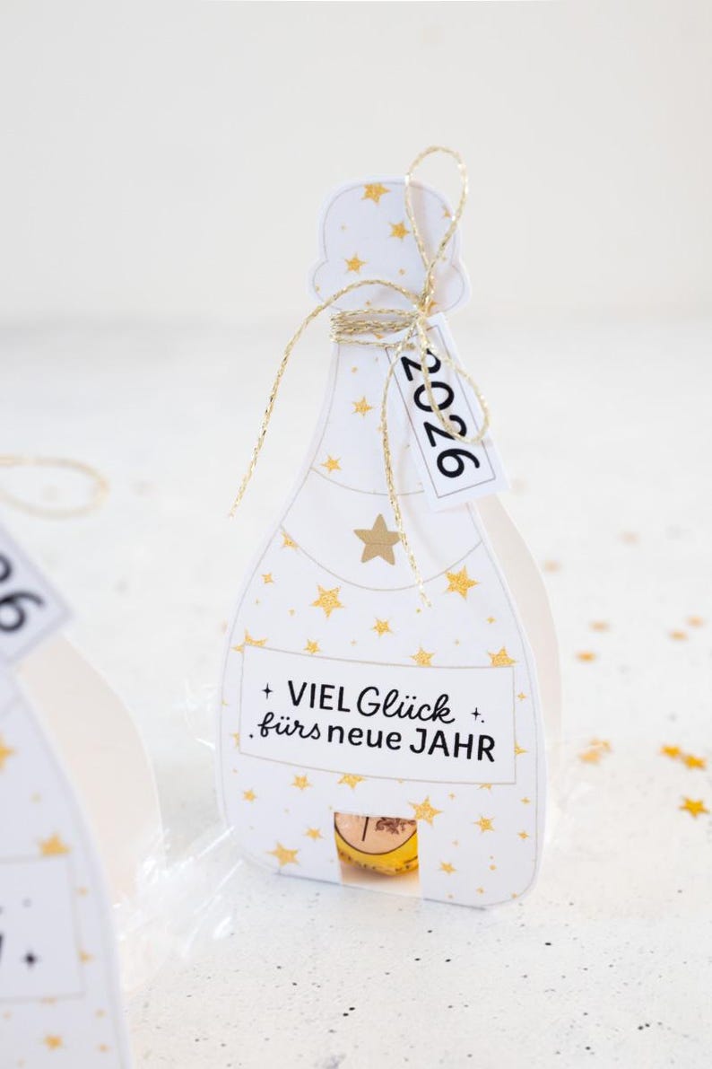 Puede incluir: Una caja de regalo blanca con forma de botella de champ&aacute;n, adornada con estrellas doradas y el texto "2026" en una etiqueta. La caja tiene una cuerda dorada y el texto alem&aacute;n "VIEL Gl&uuml;ck f&uuml;rs neue JAHR".