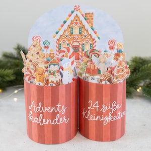 Può includere: Un calendario dell'avvento cilindrico in due parti con strisce verticali rosse. La parte anteriore presenta la scritta bianca Advents kalender e 24 süße Kleinigkeiten. La parte superiore mostra una scena pop-up con casa di pan di zenzero.