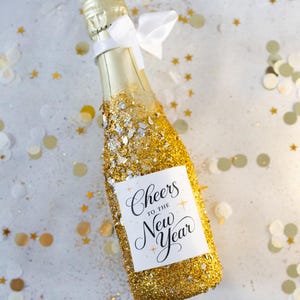 Peut inclure: Une bouteille de champagne recouverte de paillettes dorées, ornée d'un nœud blanc et d'une étiquette avec l'inscription "Cheers to the New Year". Des confettis dorés et blancs complètent la scène festive.