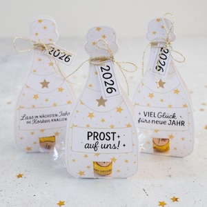 Puede incluir: Tres cajas de regalo blancas con forma de botella de champ&aacute;n decoradas con estrellas doradas. Cada caja tiene una etiqueta con "2026". Las cajas tienen texto en alem&aacute;n: "Lass im n&auml;chsten Jahr die Korken knallen", "PROST auf uns!", y "+ VIEL Gl&uuml;ck f&uuml;rs neue JAHR".