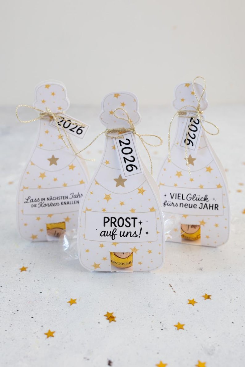 Puede incluir: Tres cajas de regalo blancas con forma de botella de champ&aacute;n decoradas con estrellas doradas. Cada caja tiene una etiqueta con "2026". Las cajas tienen texto en alem&aacute;n: "Lass im n&auml;chsten Jahr die Korken knallen", "PROST auf uns!", y "+ VIEL Gl&uuml;ck f&uuml;rs neue JAHR".
