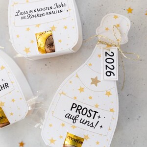 Puede incluir: Cajas de regalo blancas en forma de botella de champ&aacute;n con detalles de estrellas doradas y texto. Una caja tiene una etiqueta que dice "2026". Las cajas contienen un art&iacute;culo envuelto visible a trav&eacute;s de una peque&ntilde;a abertura. El texto "PROST auf uns!" es visible.