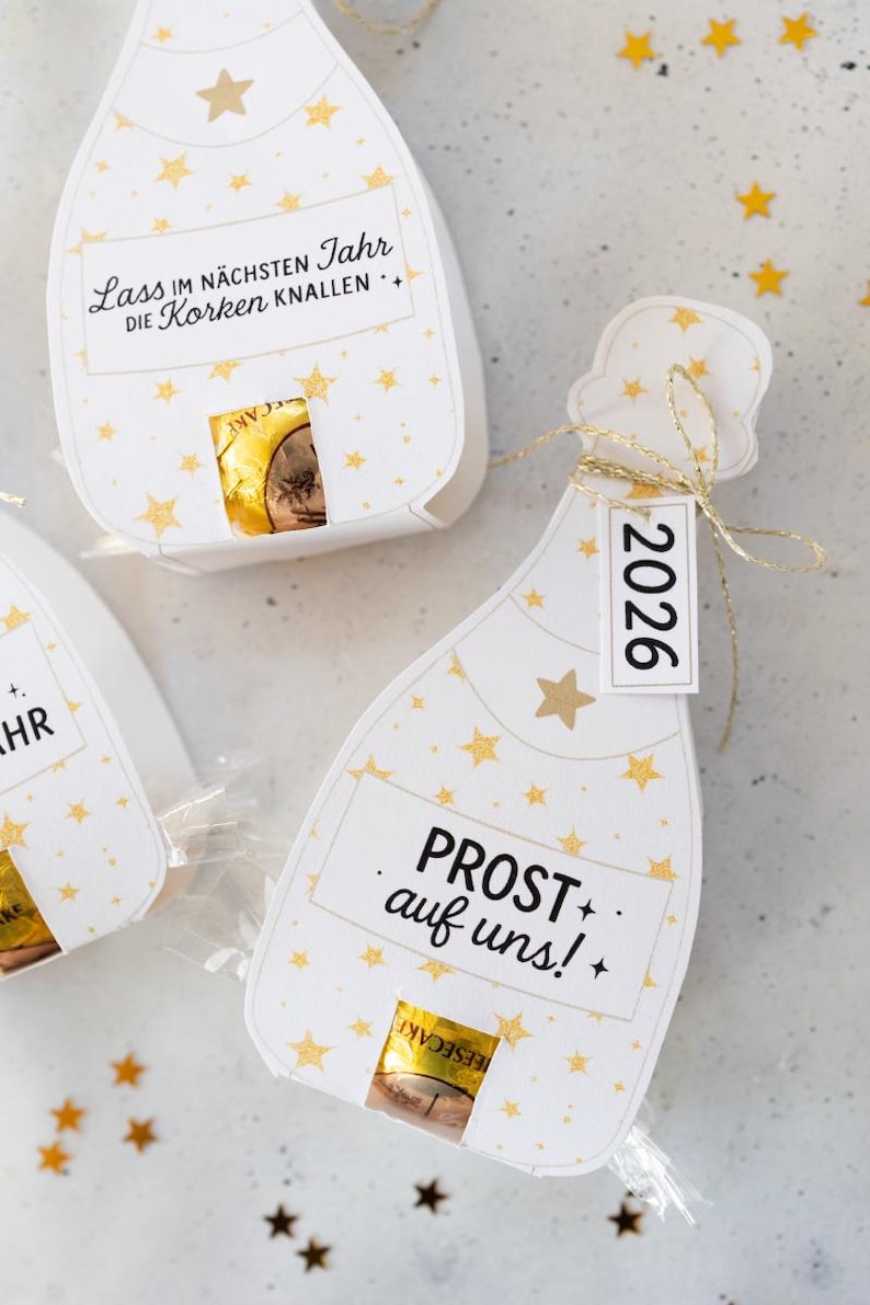 Puede incluir: Cajas de regalo blancas en forma de botella de champ&aacute;n con detalles de estrellas doradas y texto. Una caja tiene una etiqueta que dice "2026". Las cajas contienen un art&iacute;culo envuelto visible a trav&eacute;s de una peque&ntilde;a abertura. El texto "PROST auf uns!" es visible.