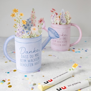 Op de afbeelding: Waterverf gieters in blauw en roze, gevuld met papieren bloemen. De blauwe kan heeft de tekst "Danke, dass du mir beim wachsen geholfen hast." Twee verpakte snoepjes met bloemaccenten zijn ook aanwezig.