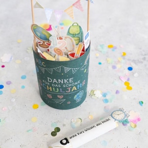 May include: A dark teal cylindrical container with school-themed stickers, a banner, and the words "Danke f&uuml;r das sch&ouml;ne Schuljahr." A white tube with the words "anke f&uuml;rs Wissen, das bleibt" is next to the container. Colorful confetti surrounds the objects.