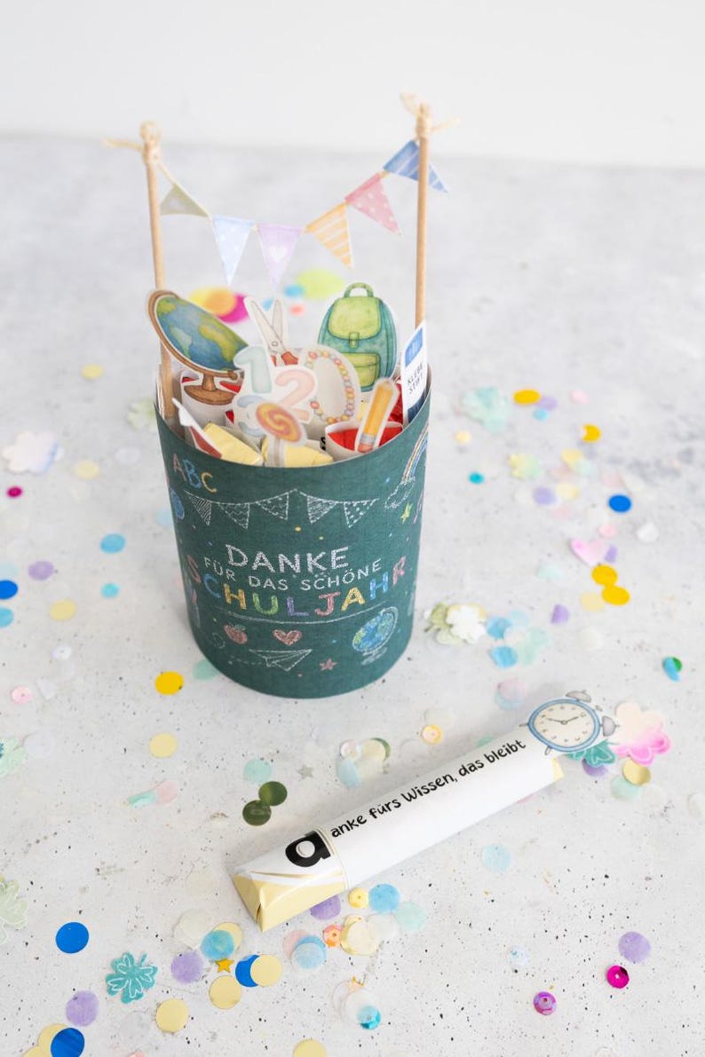 May include: A dark teal cylindrical container with school-themed stickers, a banner, and the words "Danke f&uuml;r das sch&ouml;ne Schuljahr." A white tube with the words "anke f&uuml;rs Wissen, das bleibt" is next to the container. Colorful confetti surrounds the objects.