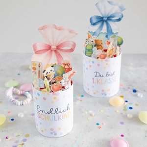 Puede incluir: Dos cajas de regalo cilíndricas con confeti colorido. Una caja tiene un lazo rosa y el texto "Endlich SCHULKIND". La otra tiene un lazo azul y el texto "Du bist 1.KLASSE". Ambas están llenas de decoraciones escolares.