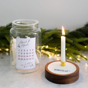Może przedstawiać: Przezroczysty szklany słoik z etykietą o treści Advent, Advent, 1-24 oraz ein Lichtlein brennt, zawierający białe świece. Obok znajduje się okrągły drewniany świecznik z białą wkładką i złotym metalowym kielichem, w którym pali się biała świeca.
