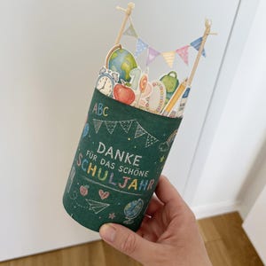 May include: A cylindrical green container with school-themed decorations and the words "DANKE F&Uuml;R DAS SCH&Ouml;NE SCHULJAHR" in white chalk-style lettering. The container is adorned with illustrations of a globe, apple, pencil, and other school supplies.