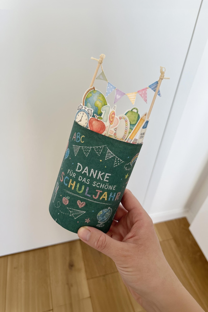 May include: A cylindrical green container with school-themed decorations and the words "DANKE F&Uuml;R DAS SCH&Ouml;NE SCHULJAHR" in white chalk-style lettering. The container is adorned with illustrations of a globe, apple, pencil, and other school supplies.